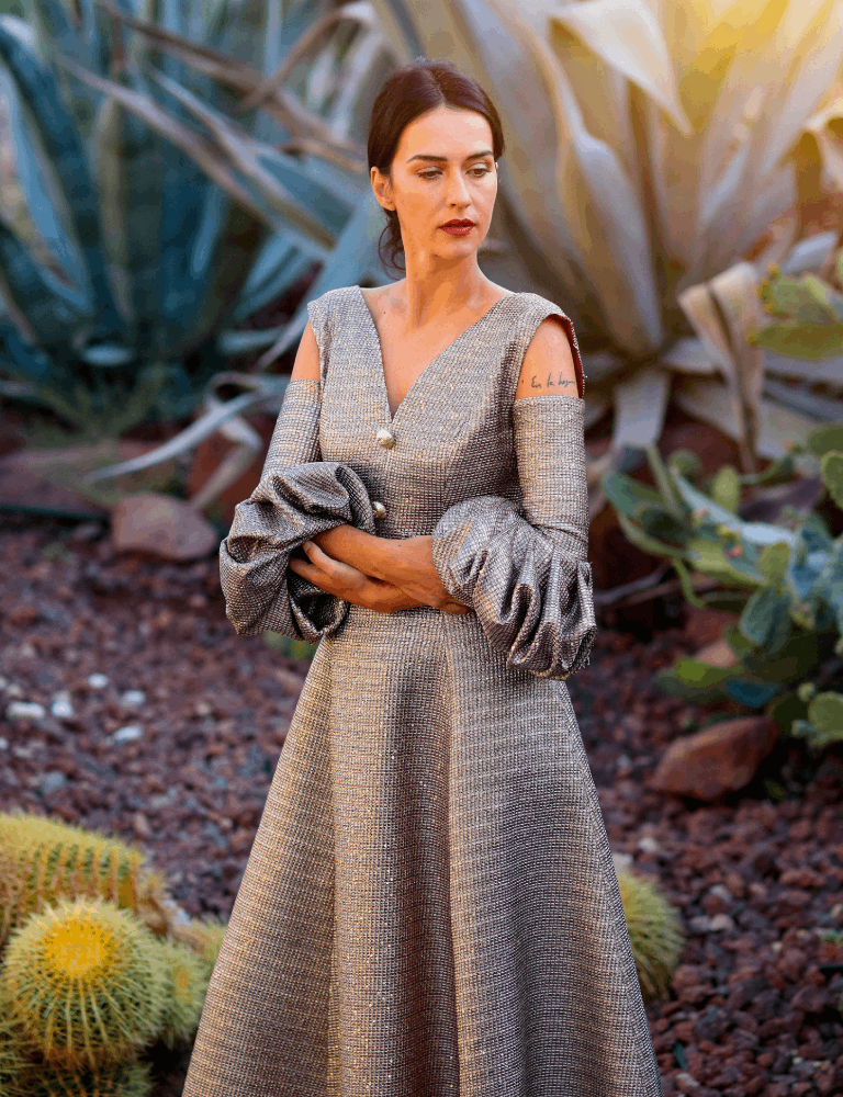 Woman wearing a metallic textured Fely Campo occasion dress with cold shoulder cutouts, voluminous puff sleeves, and a full A-line skirt, standing in a desert garden.